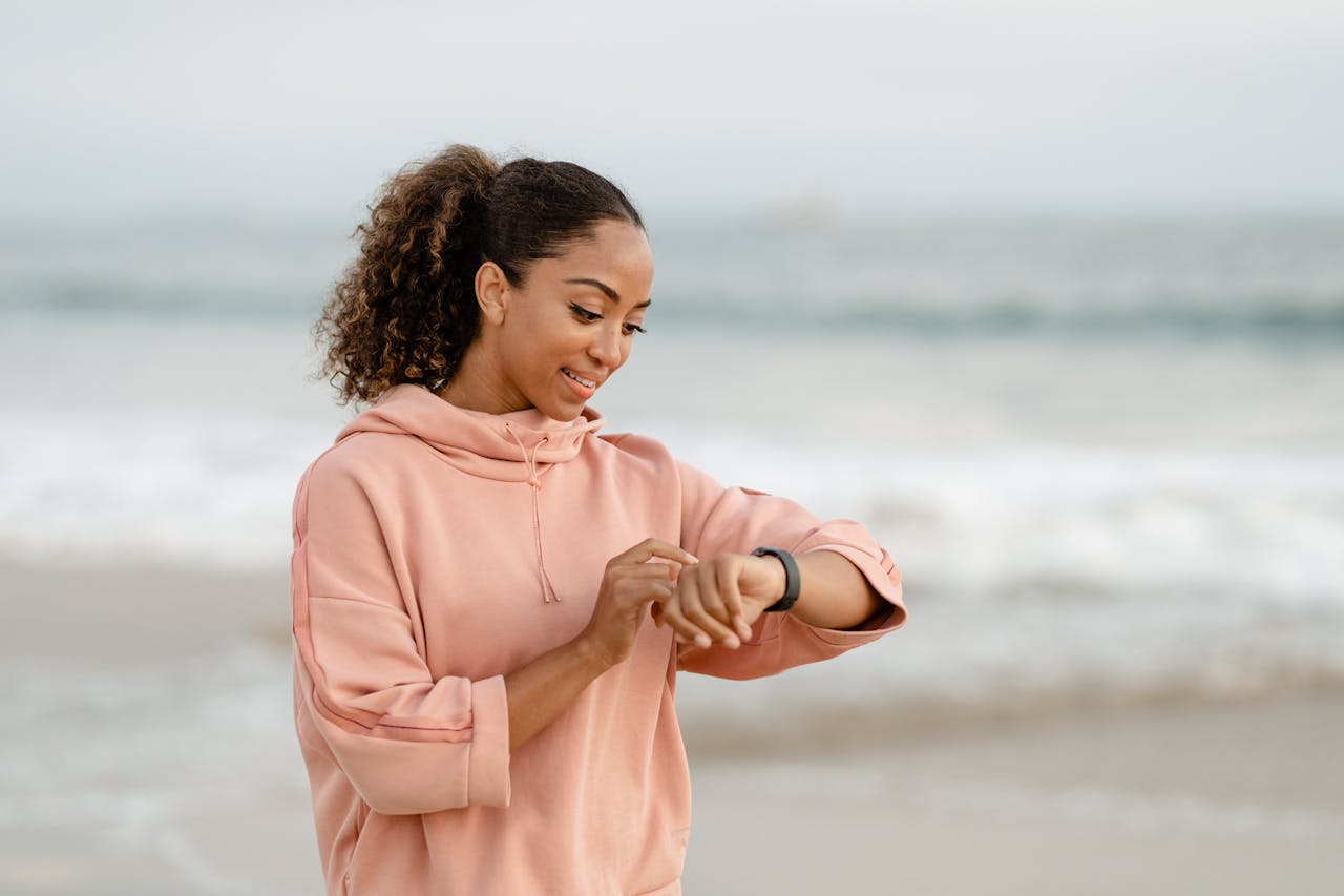 heros-img Smiling woman in a pink hoodie checks her smartwatch by the ocean.